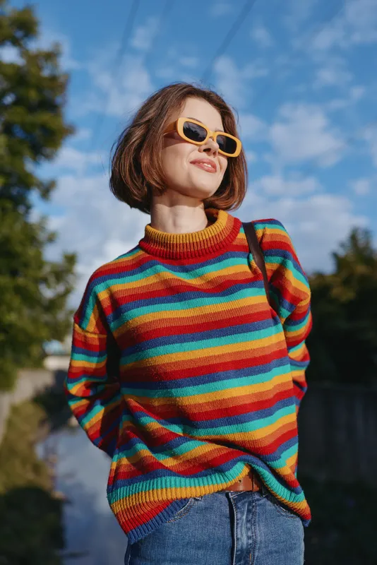 woman colorful sweater sunglasses smiling outdoor casual outfit: young woman in her 20s with bob haircut wearing a rainbow striped sweater and yellow sunglasses, smiling on a sunny outdoor path