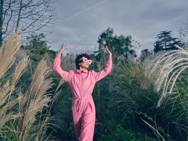 Outdoor scene with a person in a pink jumpsuit dancing among tall grasses under a blue sky, conveying freedom, joy and playful energy in nature.