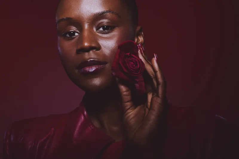 Close-up of a dark skinned African woman holding a red rose near her face against a dark red background. Concept of beauty, elegance, and natural skin glow in studio lighting.