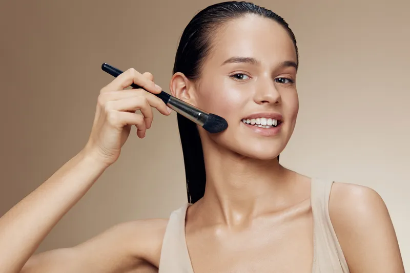 Young woman applying makeup with a brush, showcasing a natural look and glowing skin against a soft beige background, ideal for beauty and skincare concepts