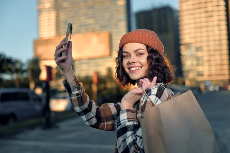 Young woman selfie with shopping bag in urban street at golden hour, candid smile and authenticity captured in golden hour glow, mindful living and emotional storytelling.