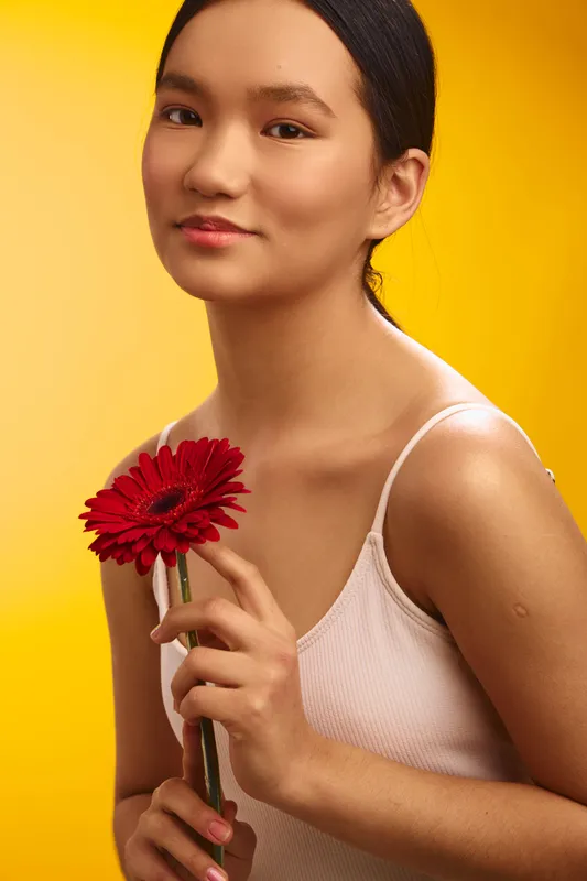Asian woman holds a vivid red flower against a vibrant yellow background, presenting a calm, graceful presence with soft lighting and minimalist studio styling.