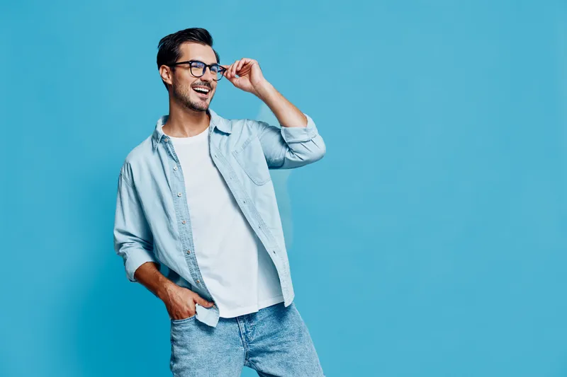 Stylish smiling man wearing glasses and denim shirt standing against a vibrant blue background, showing a cheerful expression and an approachable demeanor.