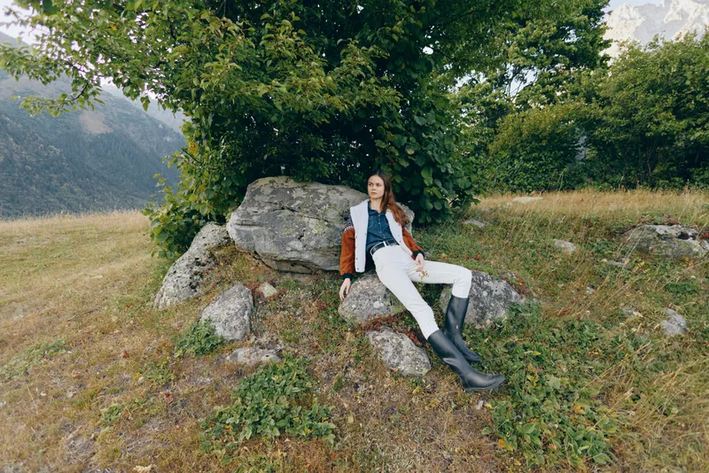 Woman outdoor in mountains meadow sitting on rocks under tree, wearing jacket and boots, relaxed pose in scenic alpine landscape for travel, nature and fashion lifestyle.