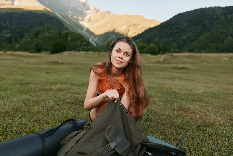 A young woman sits in a grassy field near mountains, unpacking a backpack and arranging gear. She smiles softly, enjoying an outdoor adventure in a tranquil landscape.