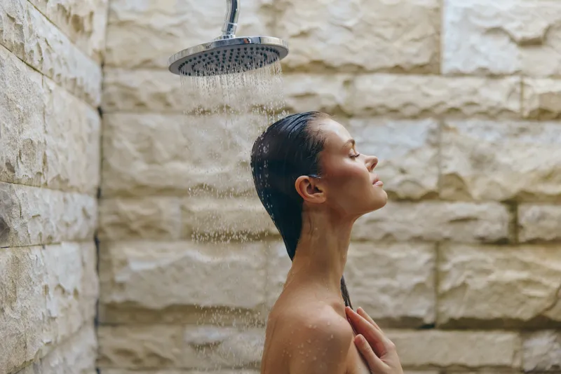 young woman enjoying outdoor shower with wet hair against stone wall, peaceful expression, natural light, wellness and relaxation concept