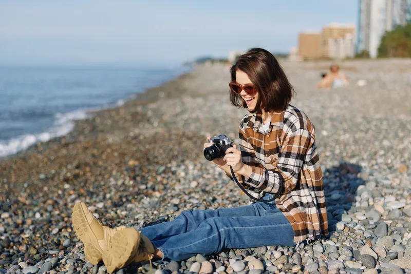 Casual woman on a pebble beach, wearing plaid shirt and jeans, sits with a camera ready for outdoor photography, enjoying sunlight, sea breeze and relaxed mood while exploring coastal scenery.