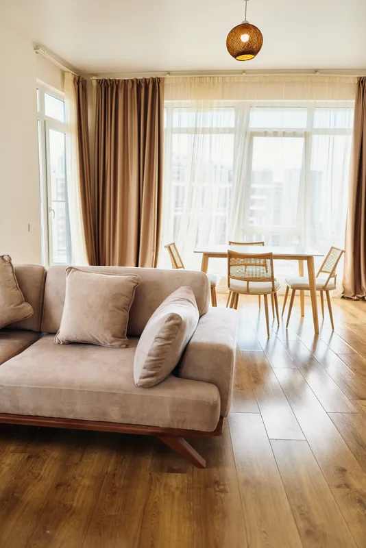 Living room with beige sofa and cushions, dining table and chairs near large windows, curtains drawn to let natural light fill warm hardwood floor in a modern cozy interior.