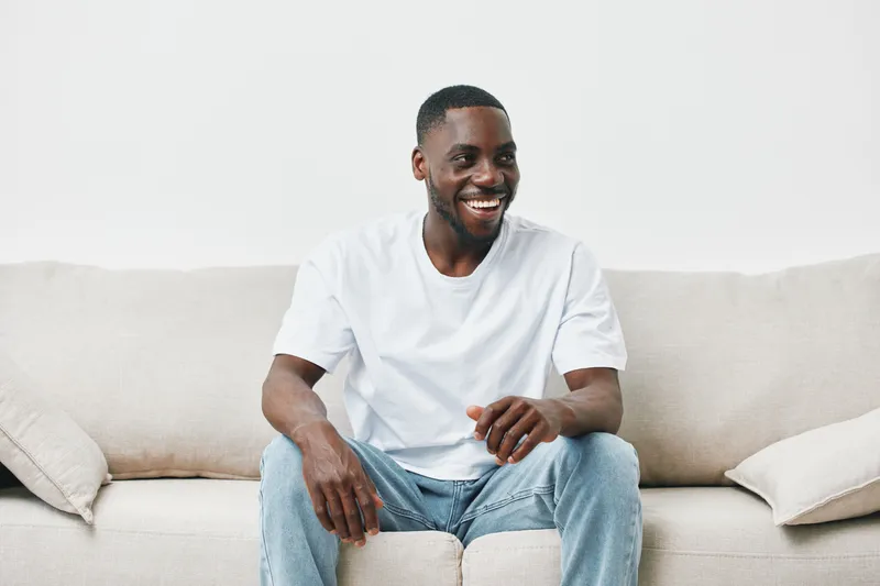 Happy young Black man sitting on a beige couch, wearing a white t shirt and jeans, embodying joy and comfort, ideal for lifestyle and wellness themes