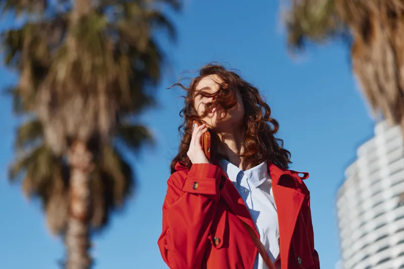 A young woman wearing a red coat stands outdoors beneath a blue sky, looking up with a gentle smile as the wind moves her hair.