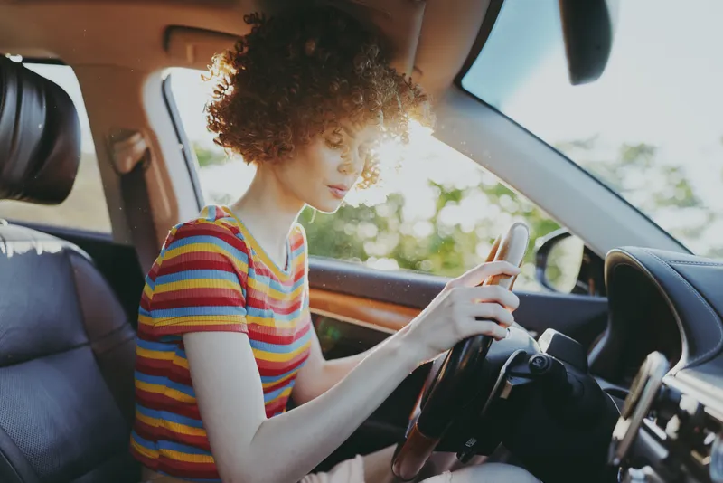 Woman driver sits in car interior with striped shirt, enjoying sunny day, retro film color mood, casual lifestyle scene, candid moment of focus and travel