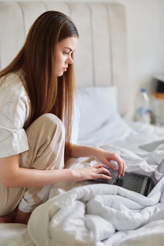 Woman on bed in bedroom working on laptop at home in pajamas, focused and typing on a small computer while sitting on a cozy duvet with soft natural light for remote work or study.