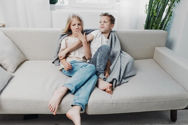 Two children sit barefoot on a beige sofa wrapped in a gray blanket, showing tired and sleepy expressions in a bright living room.
