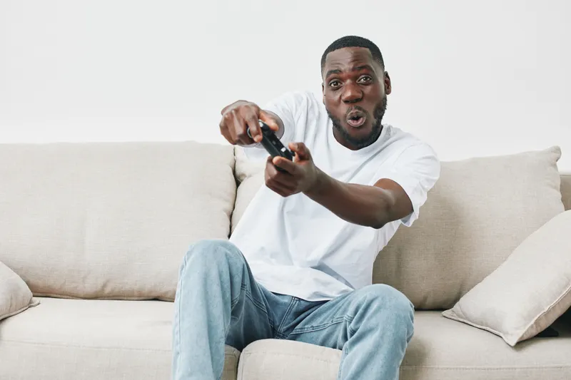 Excited young man playing video games on a couch, showing enthusiasm and joy, wearing a casual white t shirt and jeans, with a neutral background