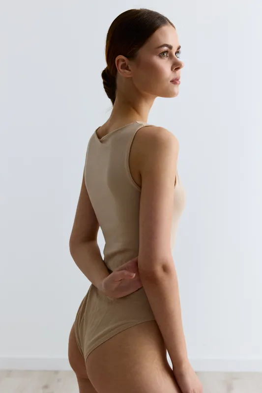 young woman in beige bodysuit standing sideways with hand behind back against plain light background, natural light highlighting smooth skin and serene expression