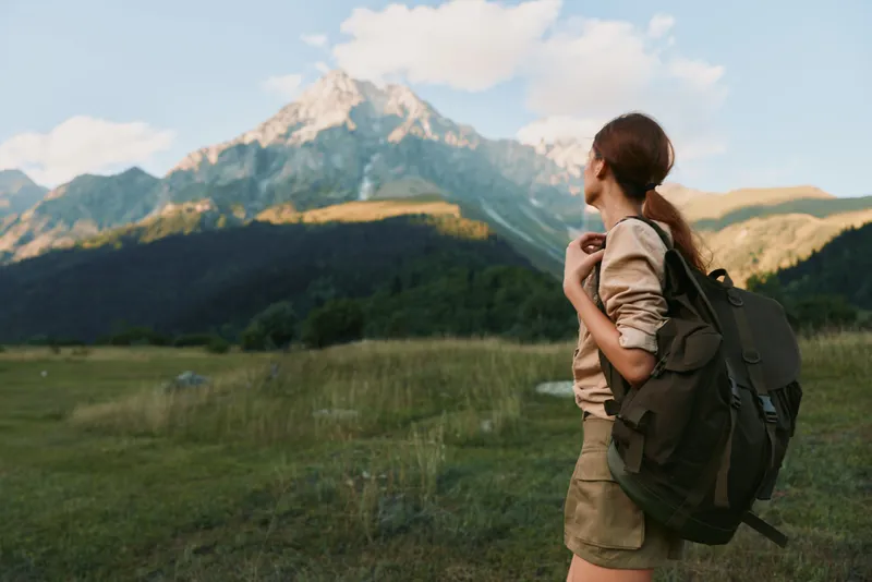 Woman with backpack standing in a grassy mountain meadow, exploring outdoors, warm light, distant peaks, casual clothing, serene mood, natural colors, and sense of adventure.