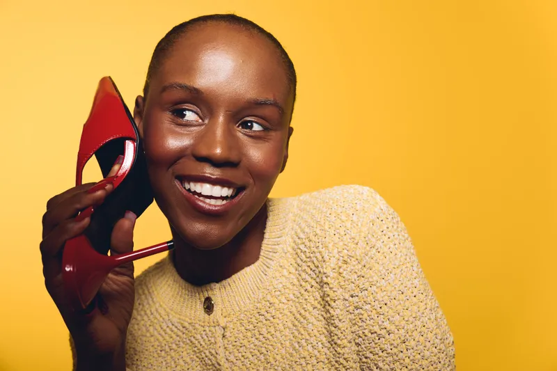 Smiling black woman with dark skin holds red high heel shoe near face against yellow background. Concept of fashion, beauty, joy, and lifestyle with natural makeup and casual clothing.