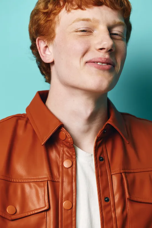 Young man with red hair wearing brown leather jacket and white t shirt posing with confident smile against blue background in studio lighting.