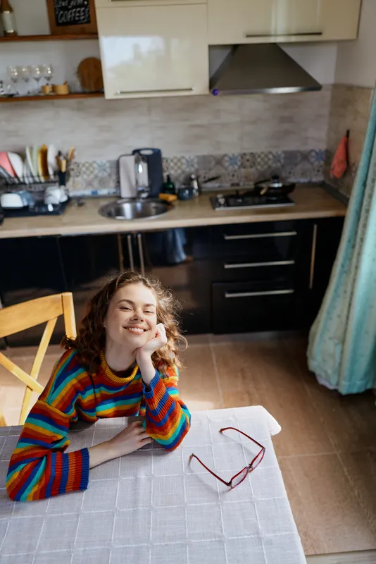 A young woman with curly hair in a colorful striped sweater rests her chin on her hand at a kitchen table, smiling softly in a cozy home moment.