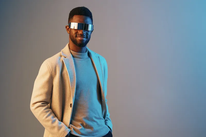 Stylish young man wearing futuristic sunglasses and a blazer, posing confidently against a blue gradient background, conveying a modern and trendy fashion concept