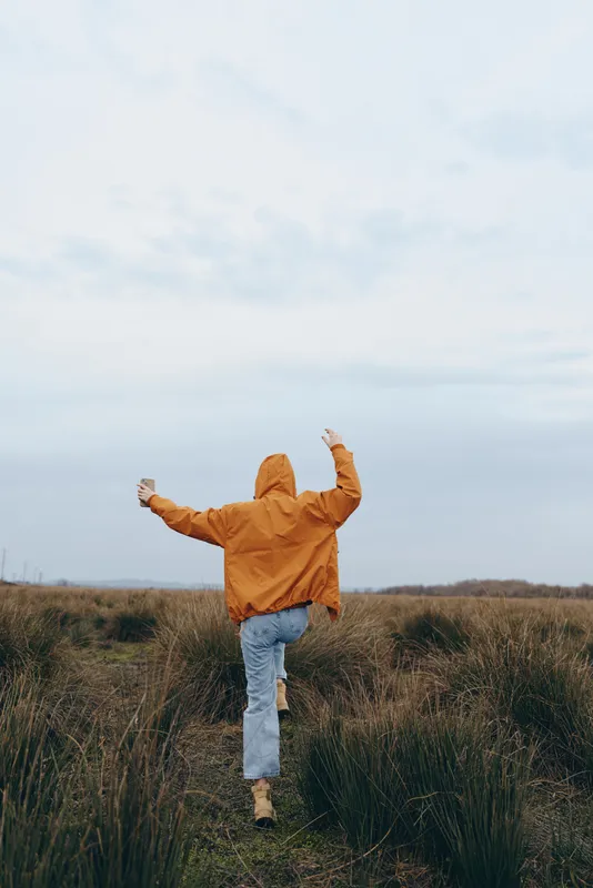 Woman outdoor walking outdoors in a field of grass, wearing an orange jacket, seen from behind against cloudy sky, nature lifestyle concept of freedom, rural calm and fresh air
