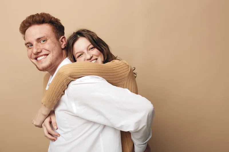 Happy young couple with red hair and brown hair smiling and hugging each other on beige background. Concept of love, friendship, relationship, and positive emotions.