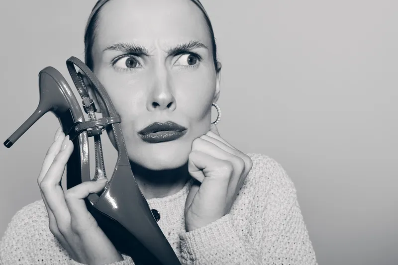 Caucasian woman with pale skin holds high heel shoe near face with confused expression on grey background. Concept for doubt, fashion, and footwear choice.