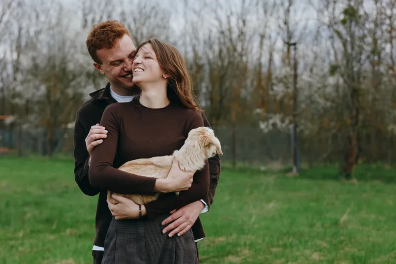 Happy couple outdoors in a park with a playful goat kid, embracing and smiling, wearing casual attire, warm light, intimate lifestyle moment, candid portrait of connection and joy