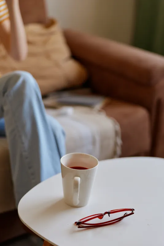 Cheerful woman enjoys coffee at home, sitting in a cozy living room with soft light, relaxed posture, warm tones, simple decor, table with cup and glasses, serene domestic scene
