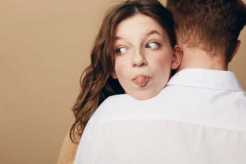 Playful young woman with brown hair sticks out tongue while hugging man in white shirt from behind. Concept of fun, affection, and casual relationship in neutral studio setting.