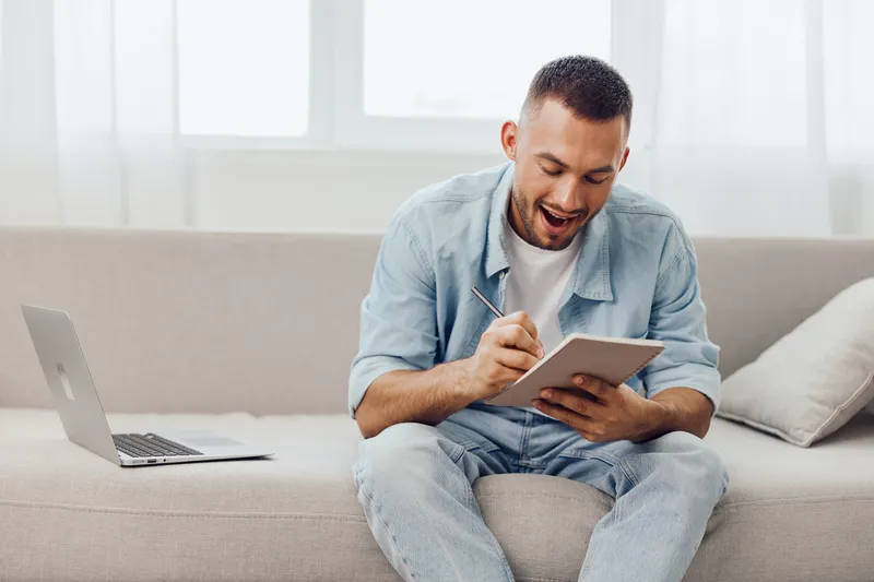 Engaged man writing notes in a cozy living room, surrounded by natural light. His relaxed smile and informal attire create a friendly, inviting atmosphere.