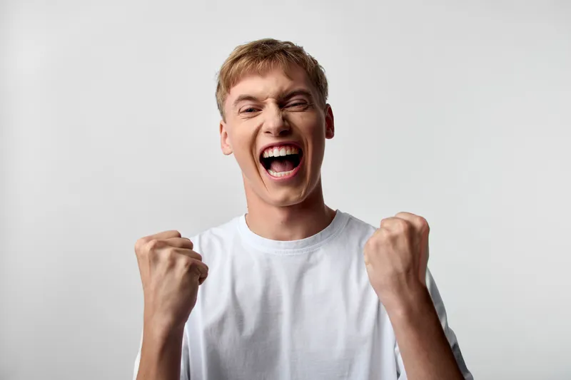Excited male with fists raised in triumph, shouting with energy and joy, capturing a dynamic moment of celebration and victory, bold expression, high adrenaline mood, plain studio backdrop, intense