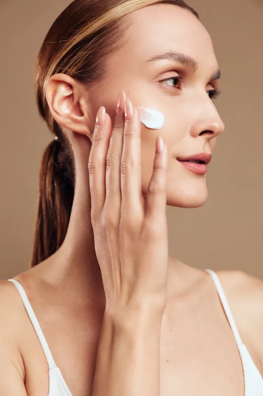Woman applying skincare cream to face, side profile close up, calm expression, gentle hand touch, soft daylight, beige studio background, beauty routine, hydration care