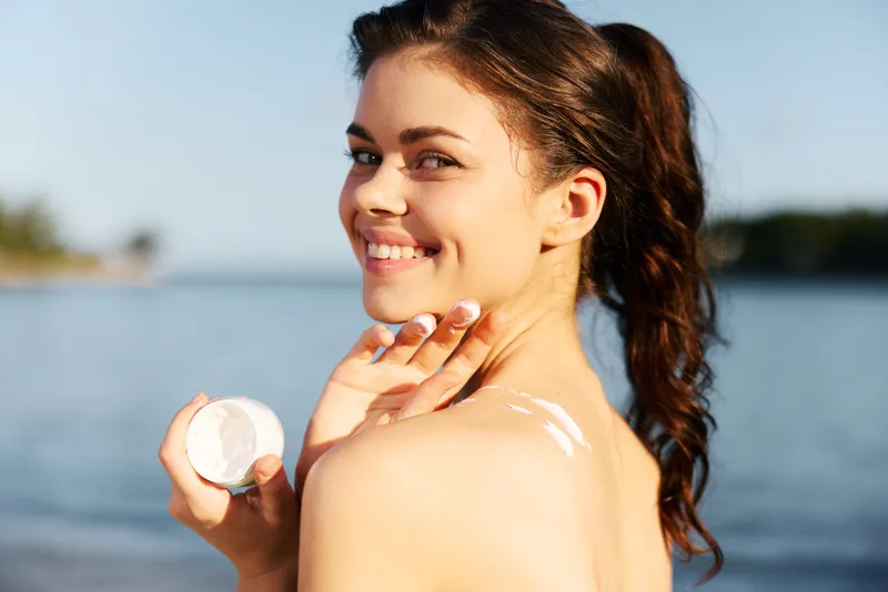 Bright portrait of a smiling woman by the water as she applies sunscreen on her shoulder, capturing sun protection, healthy skin, and a carefree outdoor moment.