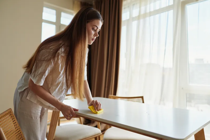 Woman cleaning dining table with yellow sponge at home, wiping surface in bright morning light by window and curtain, tidy interior with chairs and focused household chore routine.