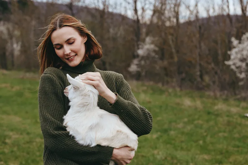 A joyful woman holds a curious goat kid in a sunlit meadow, smiling softly as they share a moment of countryside companionship and rustic lifestyle.