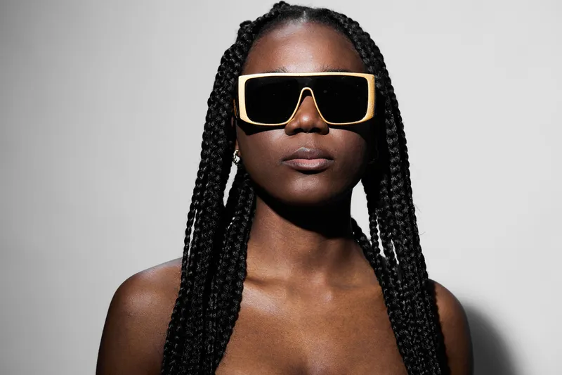 Stylish young woman with braided hair wearing oversized sunglasses, exuding confidence against a minimalist gray background, perfect for fashion themes
