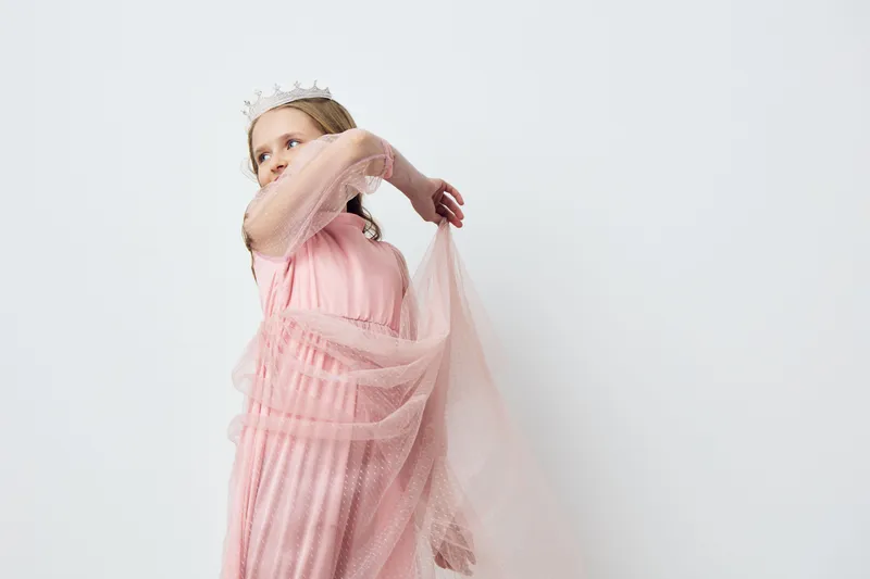 Elegant young girl in a pink dress gracefully poses with a sheer fabric, showcasing joy and innocence against a soft white background