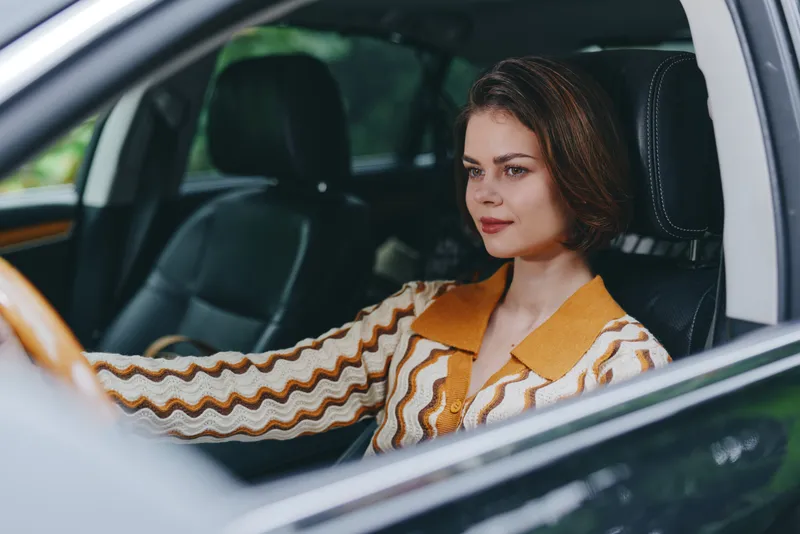 A focused woman sits behind the wheel, driving a car with calm confidence and a thoughtful expression.