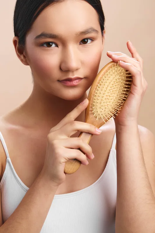 Asian woman is shown in a soft studio setting brushing her hair with a wooden brush. The calm expression and warm lighting convey a serene personal care moment.