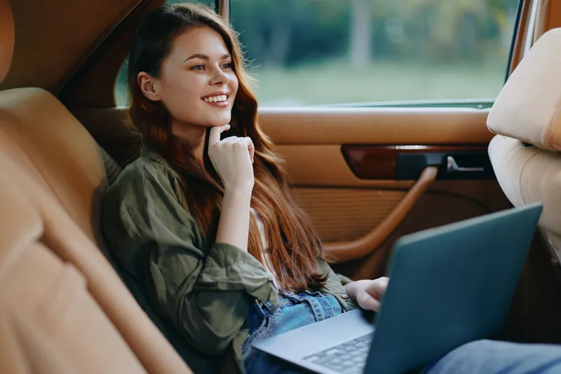 Young woman smiling while working on a laptop in a car, showcasing casual fashion and a relaxed atmosphere with natural light.