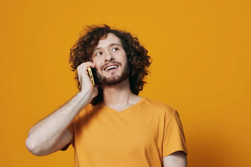 Young man with curly hair in a yellow t shirt talking on the phone against a bright yellow background, conveying happiness and vibrancy