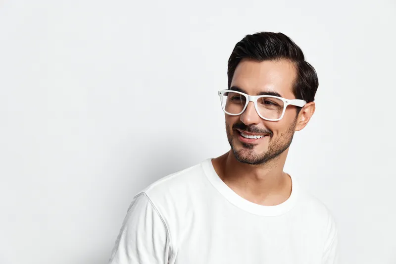 Smiling man wearing white glasses and t-shirt poses against a plain white background, showcasing a cheerful expression, capturing a moment of confidence and style.