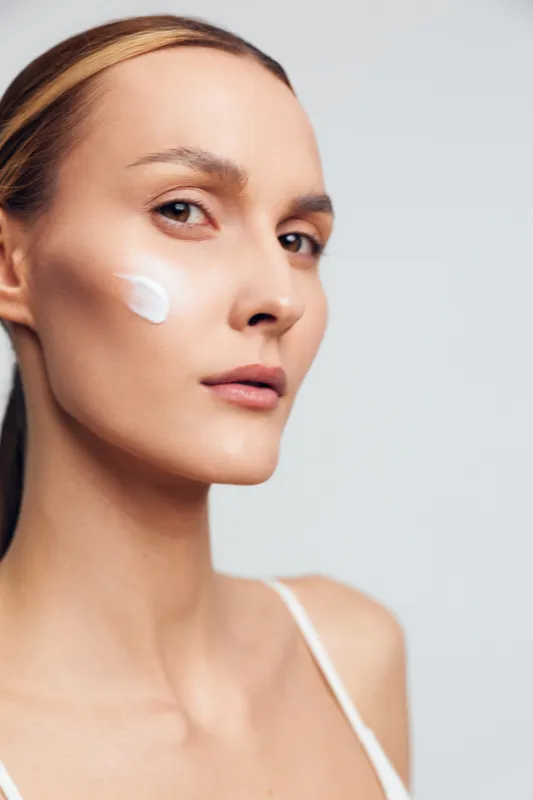 Young woman with brunette hair applies moisturizer on her cheek during a skincare routine, close up portrait with soft lighting and a neutral background