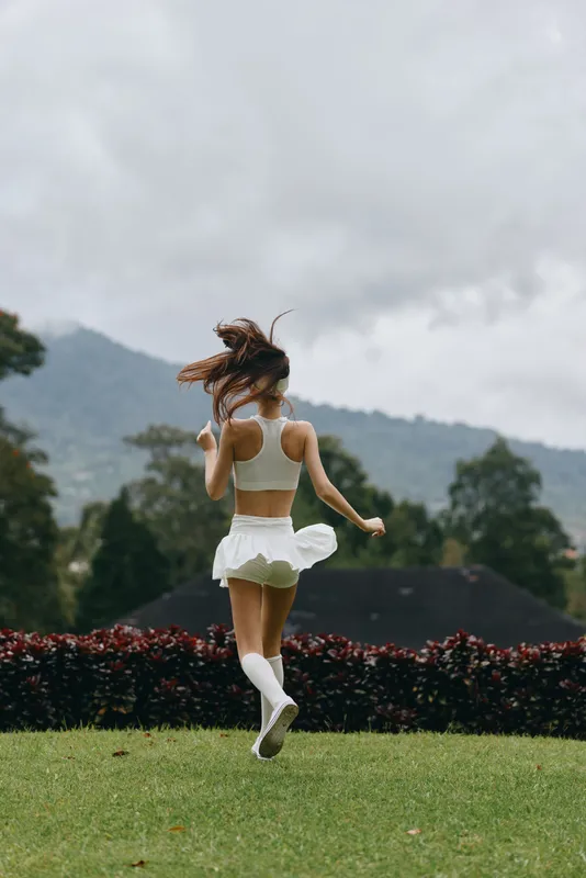 A young woman in a white athletic outfit joyfully dancing in a green meadow, surrounded by lush nature and mountains, embodying freedom and happiness