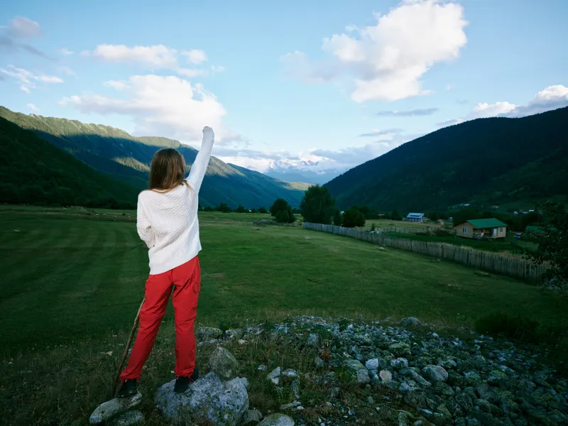 Woman standing on a rocky overlook in open countryside, arms raised toward distant mountains and valley, clear blue sky, vibrant landscape, feeling freedom and exploration