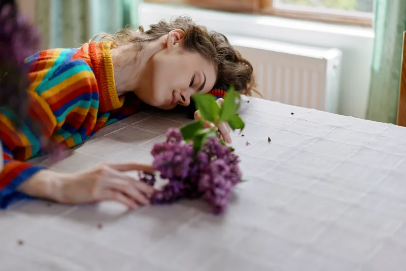 A tired woman rests her head on a table in a sunlit home, beside a bouquet of purple flowers. A simple still life exploring fatigue, rest, and mental health in a cozy domestic setting.