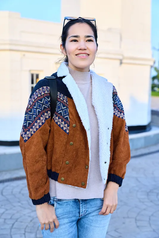 Woman in a textured brown and patterned jacket, denim jeans, outdoors on a sunny day, casual fashion style in an urban setting, candid and confident lifestyle and travel catalogs