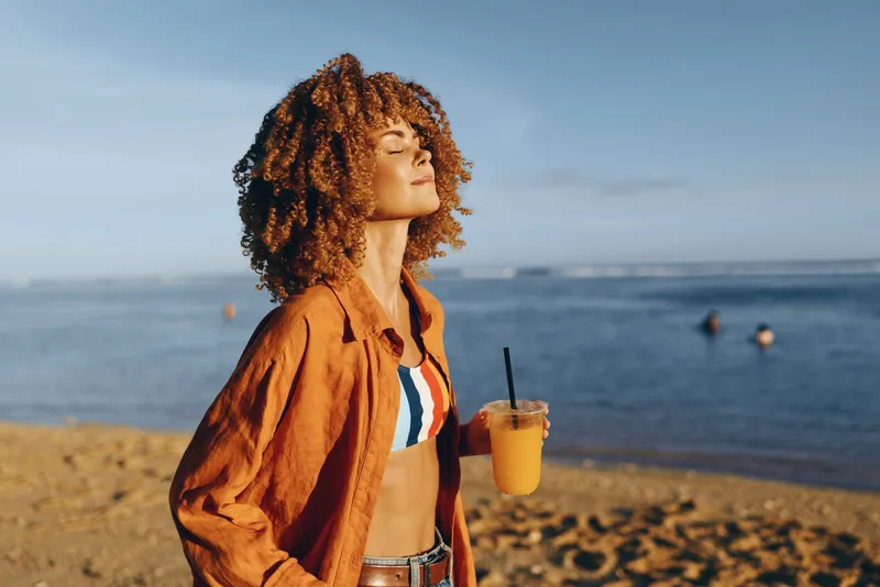 Woman, curly hair, beach, ocean, portrait, holding drink, warm sunlight, golden hour, profile pose, relaxed posture, sandy shore, ocean waves, soft breeze, blue sky