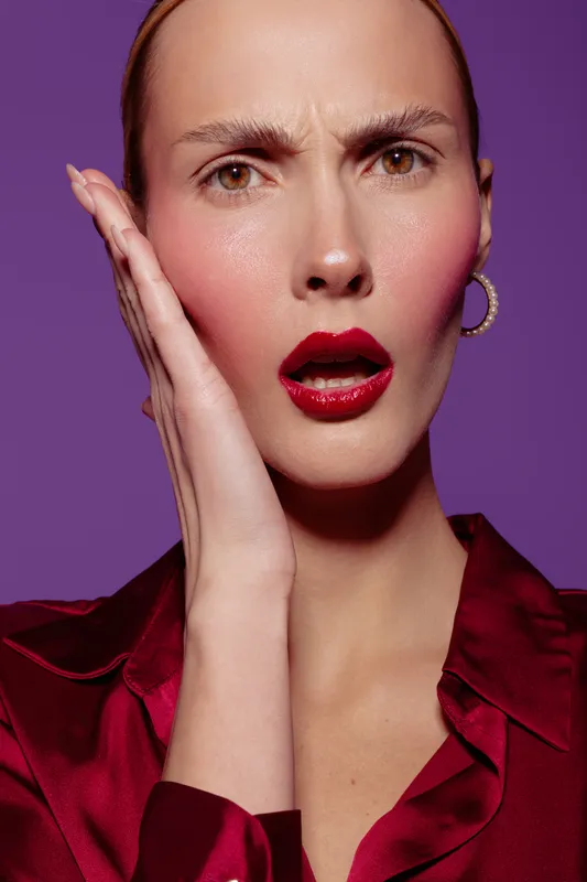 Close-up of surprised woman with red lips and hoop earrings touching her face on purple background. Concept of emotion, beauty, fashion, and makeup in studio lighting.